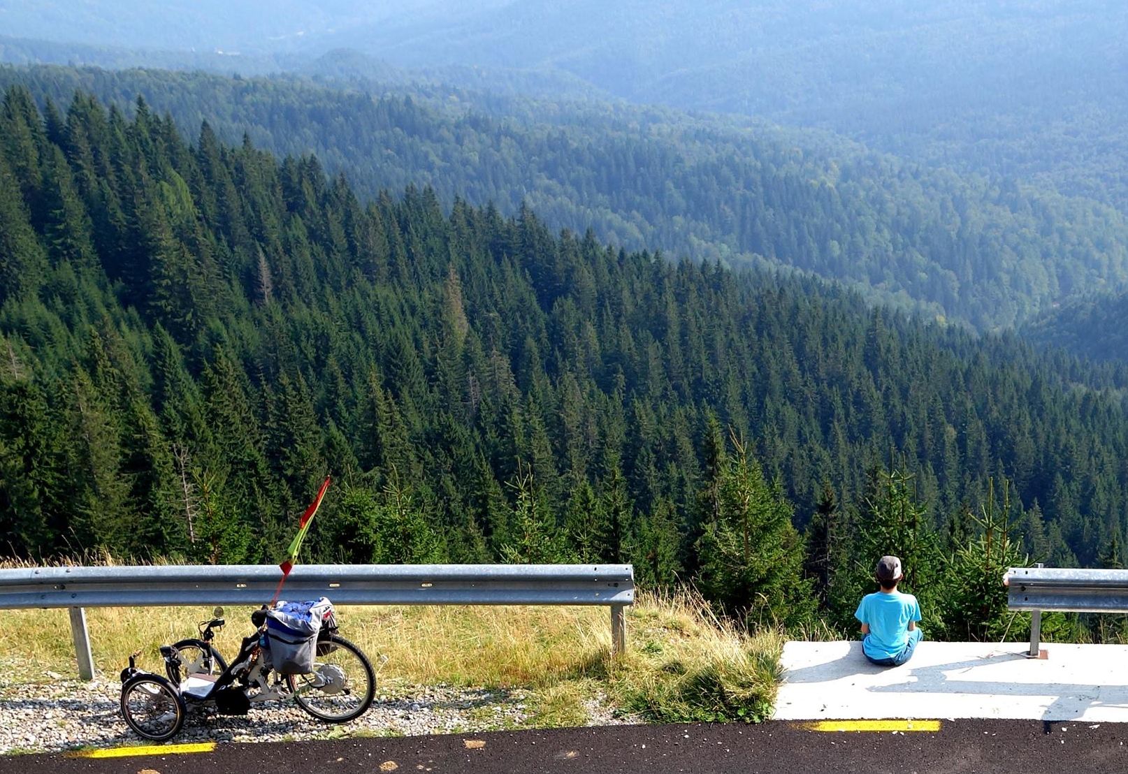 A man sits at the edge of a suspended highway overlooking forests. A trike is parked close to him. 